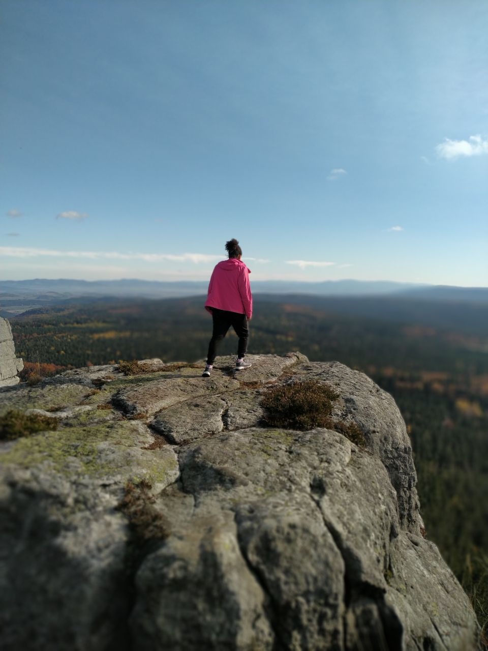 Kobieta w różowej kurtce stoi na skalistym szczycie, podziwiając rozległy krajobraz zalesionych wzgórz i błękitne niebo.