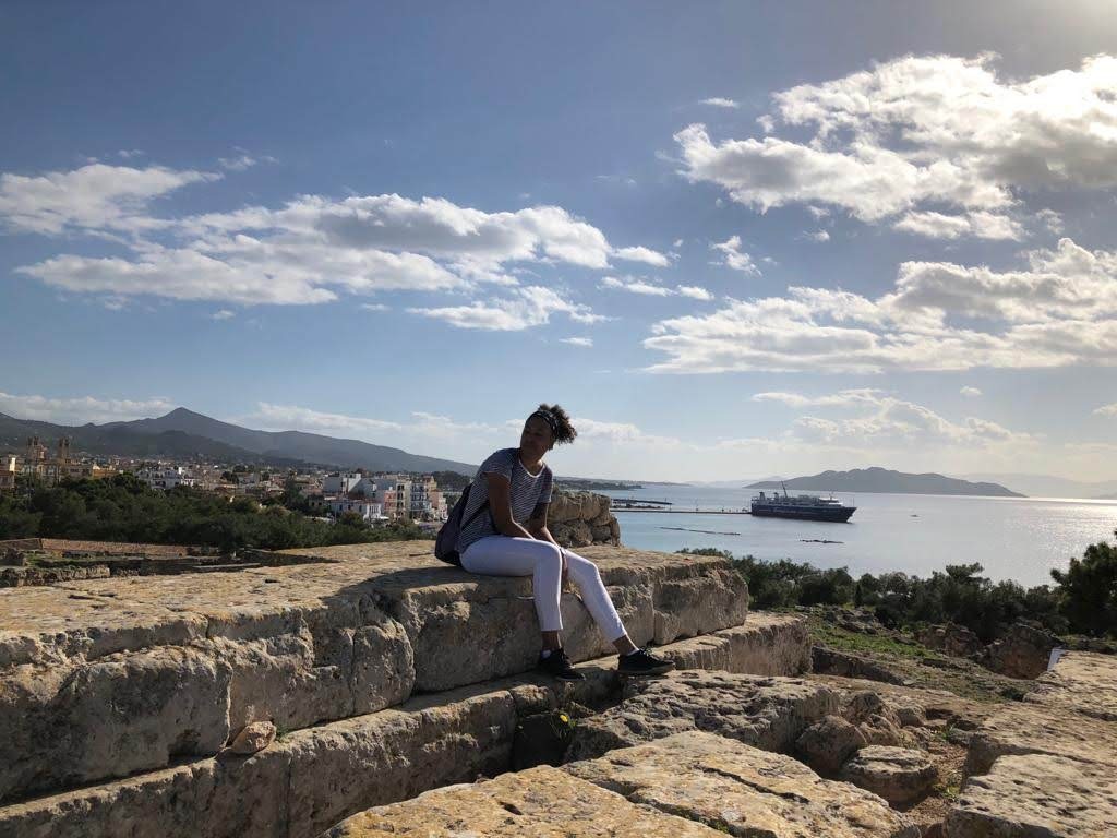 Kobieta siedzi na kamiennym murze z widokiem na miasto, morze i statek wycieczkowy w oddali, pod błękitnym niebem z białymi chmurami.
