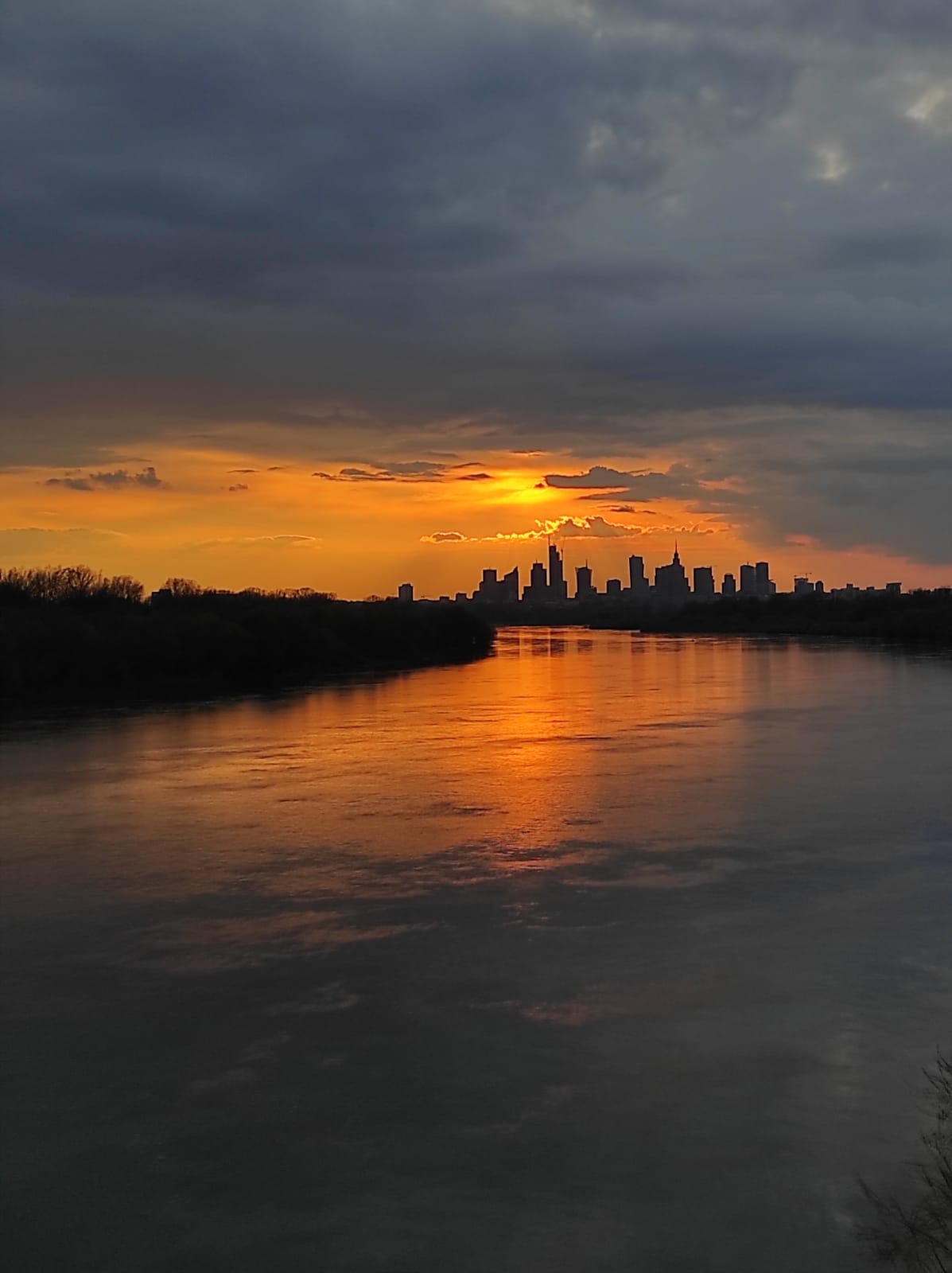 Panorama miasta w promieniach zachodzącego słońca odbijająca się w spokojnej rzece, ciemne chmury nad sylwetką wieżowców.