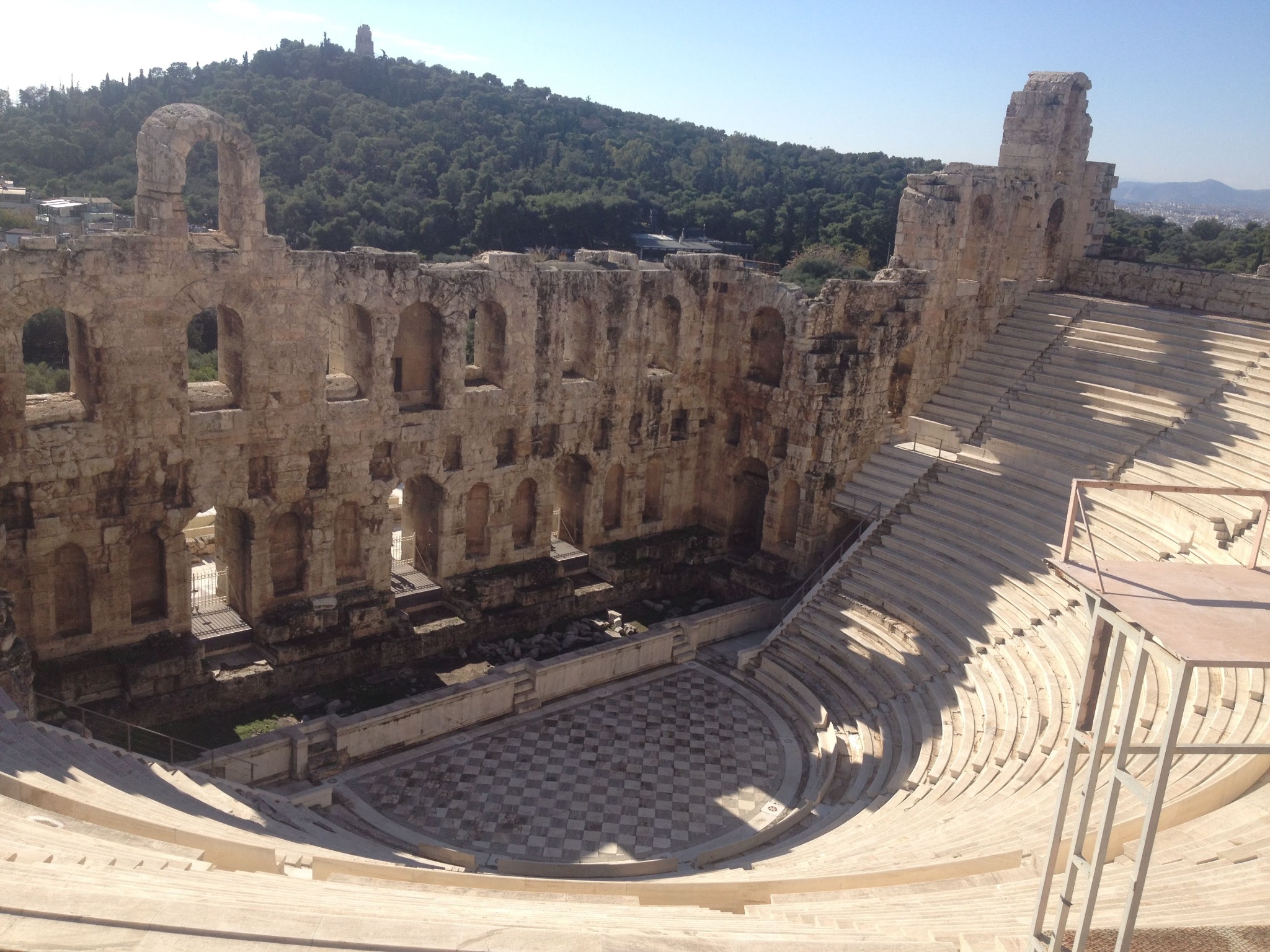 Odeon Herodesa Attykusa w Atenach, widok z góry na kamienne rzędy siedzeń i scenę z szachownicą, w tle zalesione wzgórze i błękitne niebo.