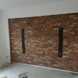 Majster - Ściana z czerwonej cegły z dwiema pionowymi, czarnymi wnękami na instalacje elektryczne w trakcie prac wykończeniowych. Na podłodze widoczne resztki materiałów budowlanych.