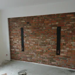 Ściana z czerwonej cegły z dwiema pionowymi, czarnymi wnękami na instalacje elektryczne w trakcie prac wykończeniowych. Na podłodze widoczne resztki materiałów budowlanych.