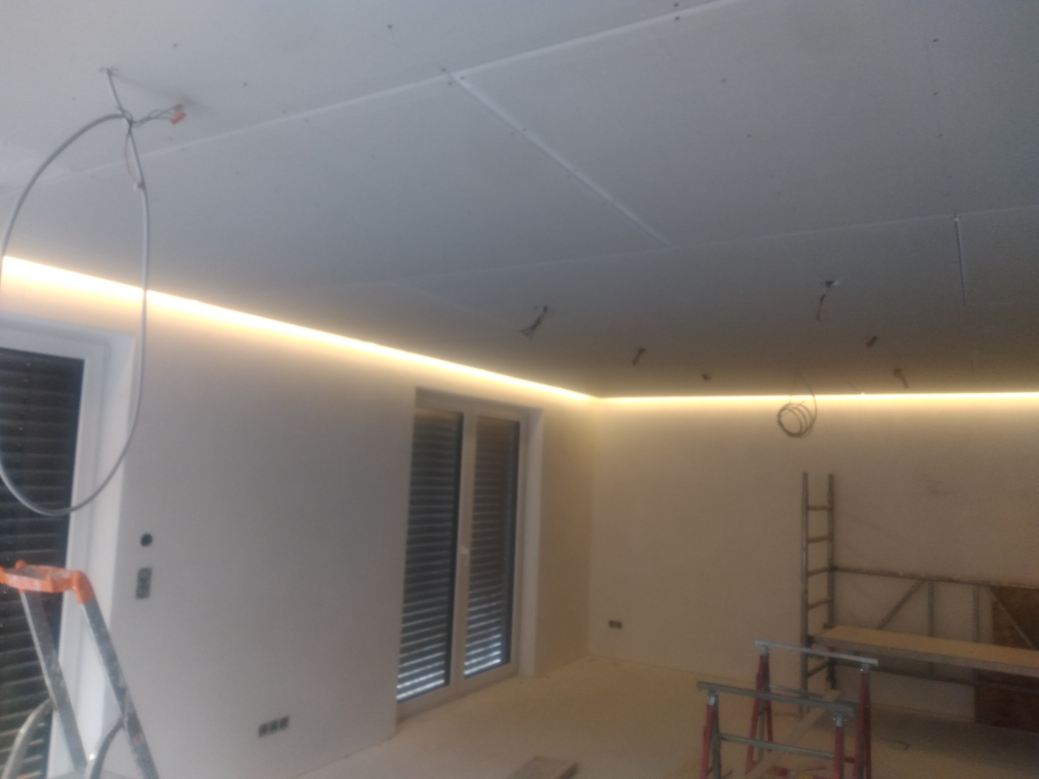 Wnętrze w trakcie prac wykończeniowych: sufit z płyt gipsowo-kartonowych ze świecącą listwą LED, widoczne przewody elektryczne, drabina i kozły malarskie.