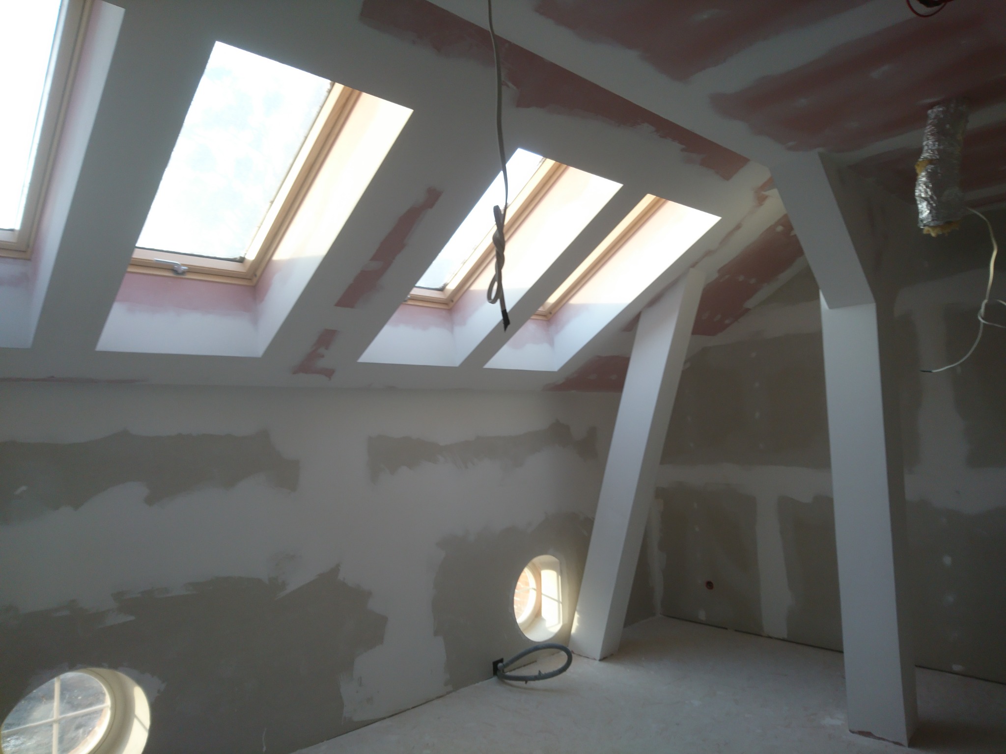 Poddasze w trakcie prac wykończeniowych w Bukowcu, widoczne okna dachowe, ściany z widocznymi śladami szpachlowania i przygotowania do malowania, okrągłe okienko w ścianie, oraz wystające kable.