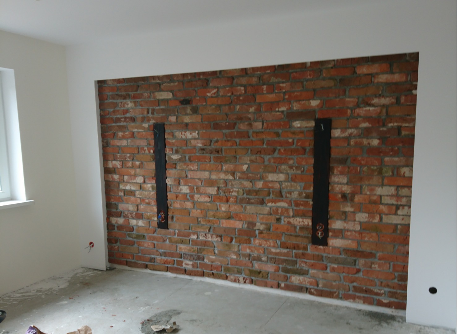 Ściana z czerwonej cegły z dwiema pionowymi, czarnymi wnękami na instalacje elektryczne w trakcie prac wykończeniowych. Na podłodze widoczne resztki materiałów budowlanych.