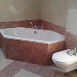 Kąpielowa wanna narożna z obudową wyłożoną brązowymi płytkami, widoczny bidet i mozaika na ścianach