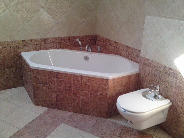 Kąpielowa wanna narożna z obudową wyłożoną brązowymi płytkami, widoczny bidet i mozaika na ścianach