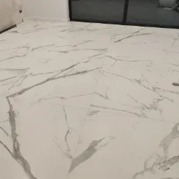 Ułożone białe płytki podłogowe imitujące marmur z szarymi żyłkami, widoczne fragmenty ściany w stanie surowym i okna balkonowego.