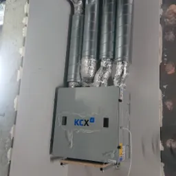 Szara centrala wentylacyjna marki KCX z czterema podłączonymi, izolowanymi przewodami wentylacyjnymi w Gdyni, zamontowana na ścianie, jeszcze częściowo zabezpieczona folią ochronną.