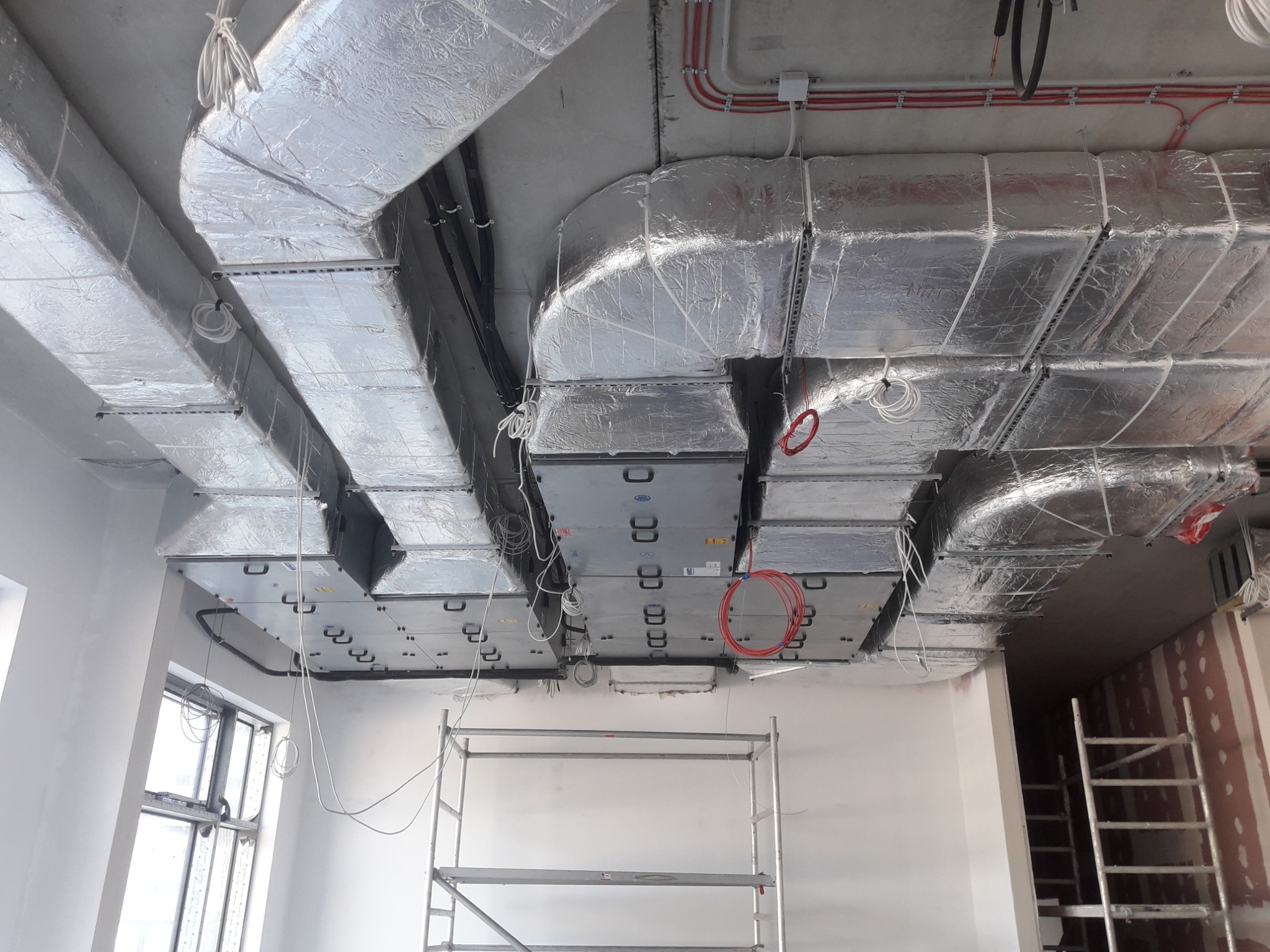 Widok na sufit z zamontowanymi kanałami wentylacyjnymi w srebrnej izolacji, widoczne także elementy instalacji elektrycznej i fragment rusztowania.