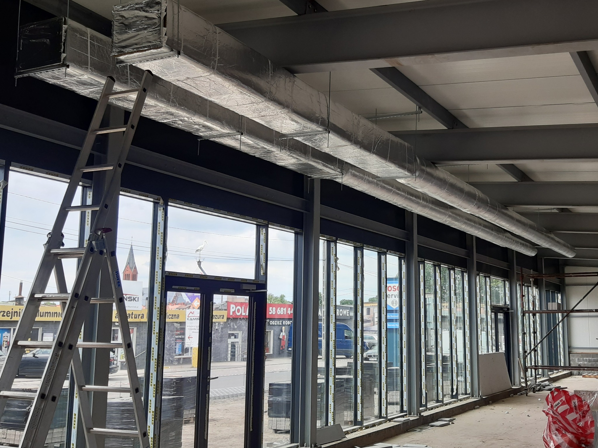 Wnętrze hali w trakcie prac instalacyjnych, widoczne srebrne, izolowane kanały wentylacyjne pod sufitem, drabina oparta o okno z widokiem na ulicę z kościołem w tle.