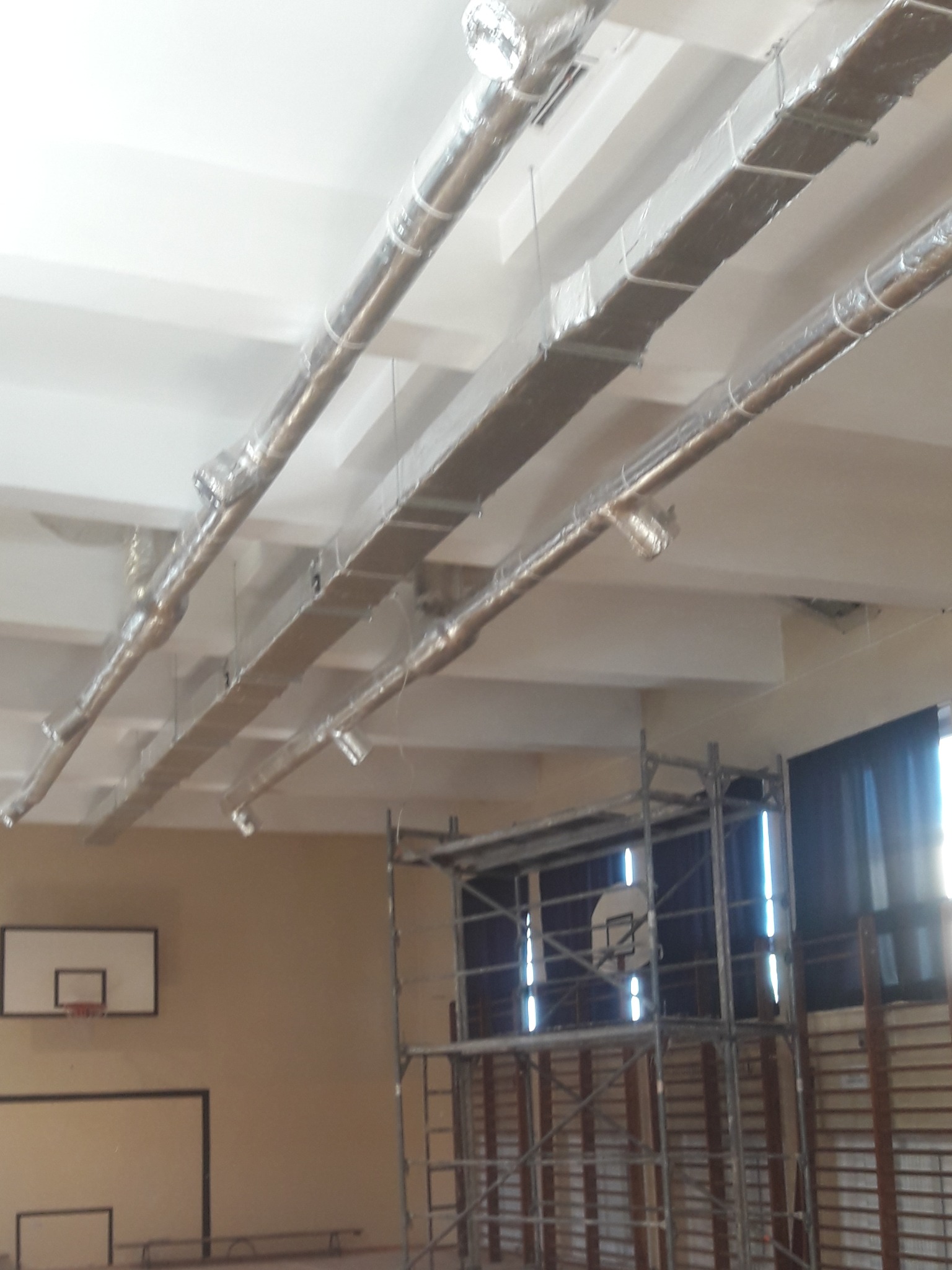 Montaż srebrnych kanałów wentylacyjnych pod sufitem w hali sportowej, widoczne rusztowanie i kosz do koszykówki.