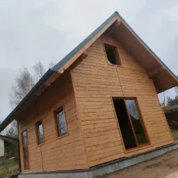 Surowy, drewniany dom szkieletowy z ciemną stolarką okienną i blachodachówką, sfotografowany pod kątem na nierównym terenie w pochmurny dzień.