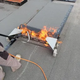 Pracownik z bliska, w roboczych spodniach i białych butach, używa specjalistycznego wózka z palnikiem do zgrzewania papy dachowej marki Lemar na płaskim dachu, obok komina z cegły. Ogień intensywny...