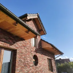 Nowoczesny dom z czerwonej cegły z okrągłym oknem, drewnianym podbitką i czarnymi rynnami na tle błękitnego nieba.