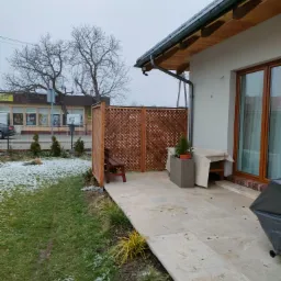 Zewnętrzny taras domu z drewnianą kratownicą, częściowo przykrytym stołem i roślinami w donicach, widok na trawnik z resztkami śniegu i budynek w tle.