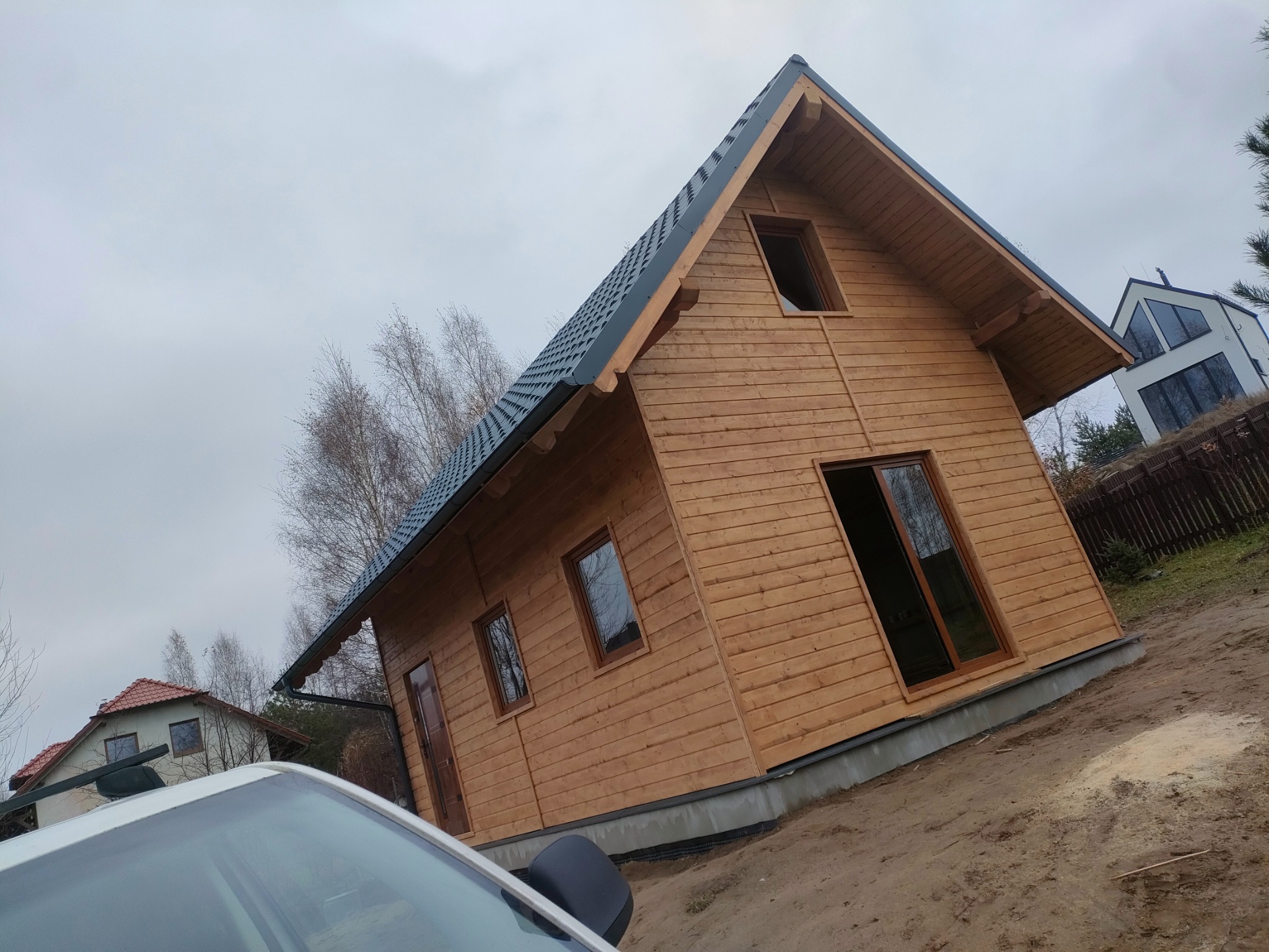 Surowy, drewniany dom w stylu skandynawskim z ciemną dachówką, widoczny z perspektywy ziemi, z fragmentem samochodu w kadrze i innymi domami w tle.