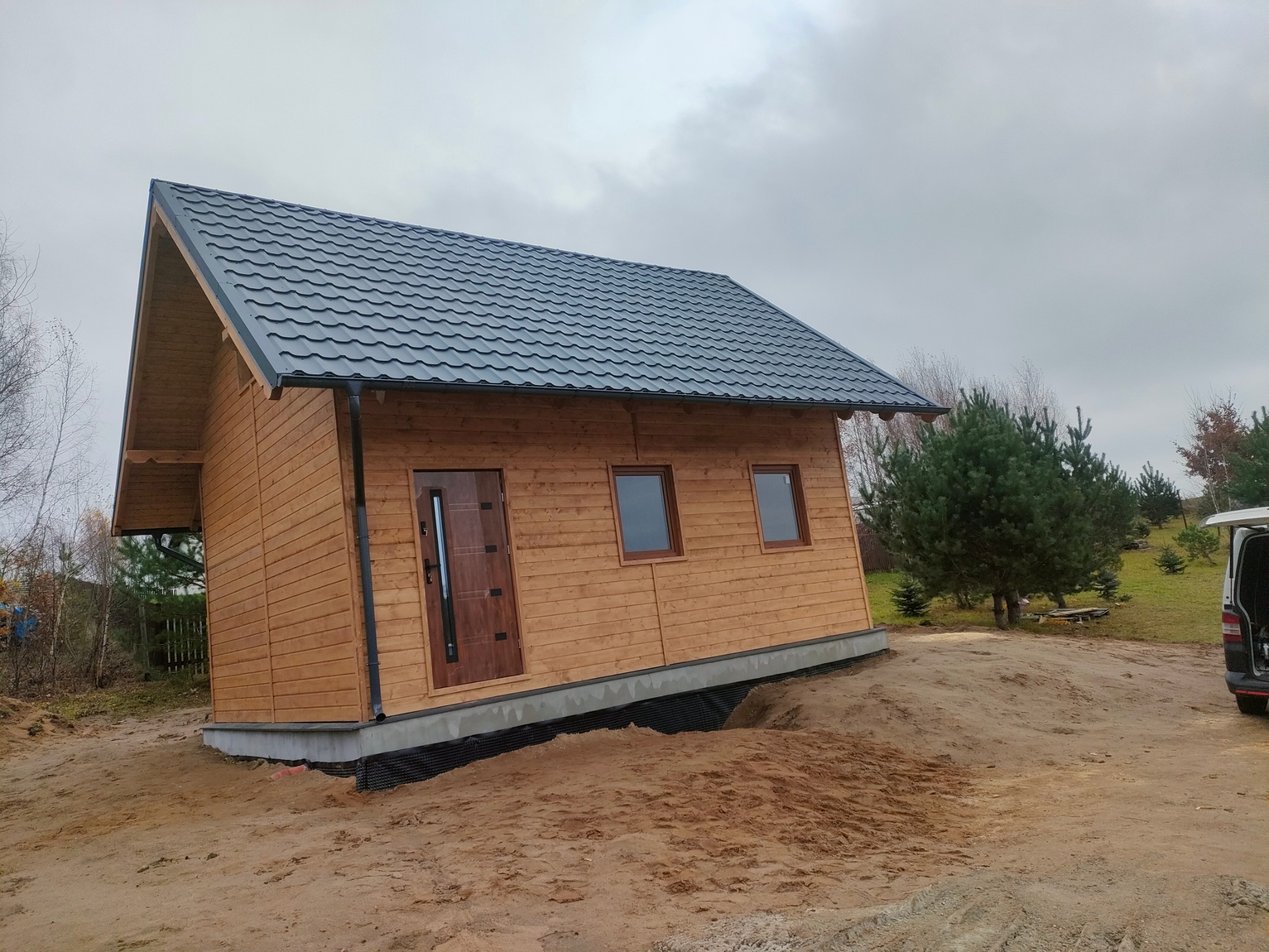 Nieduży, drewniany dom z ciemną, metalową dachówką, osadzony na betonowej podmurówce z widoczną izolacją, na piaszczystym terenie z pojedynczymi drzewami w tle.