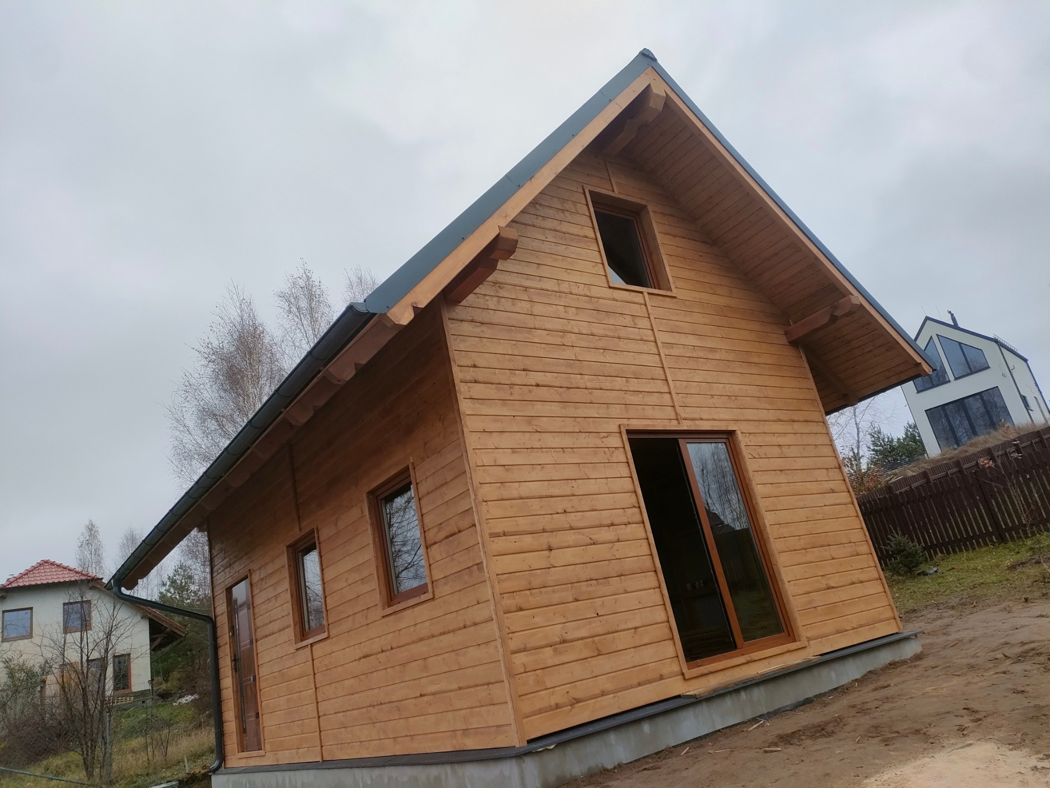 Surowy, drewniany dom szkieletowy z ciemną stolarką okienną i blachodachówką, sfotografowany pod kątem na nierównym terenie w pochmurny dzień.