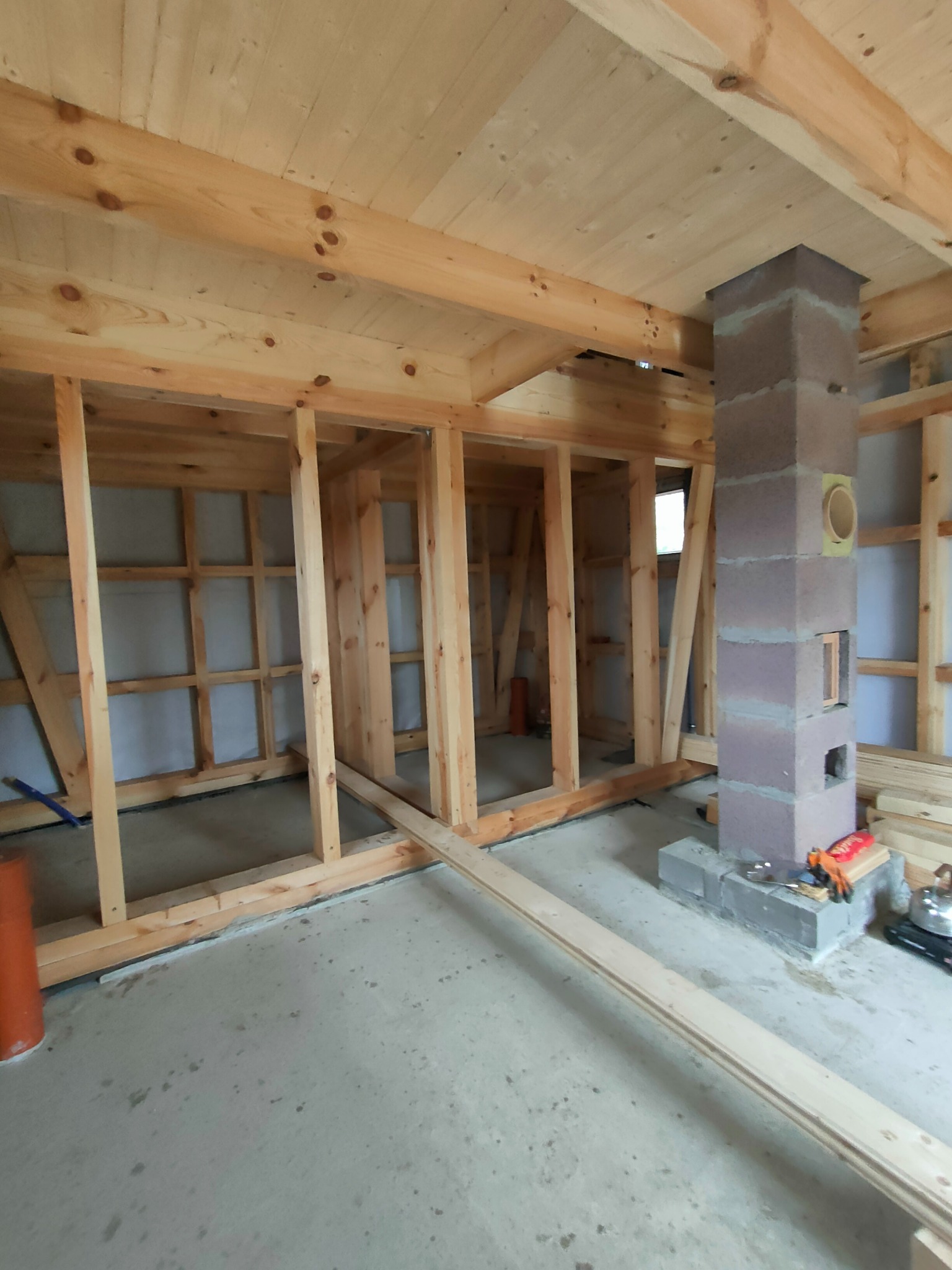 Wnętrze w trakcie budowy domu szkieletowego z widoczną konstrukcją ścian, drewnianym stropem i kominem z pustaków betonowych. Na podłodze leżą deski i narzędzia.