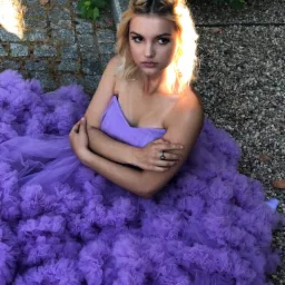 Portret kobiety w fioletowej sukni z falbanami, siedzącej na żwirowym podłożu, oświetlonej słońcem, z delikatnym makijażem i upiętymi włosami.