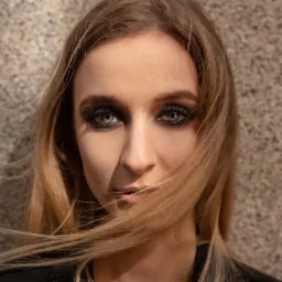 Portret młodej kobiety z intensywnym makijażem oczu w stylu smoky eye, z blond włosami delikatnie rozwianymi na twarzy, na tle szarej, teksturowanej ściany.