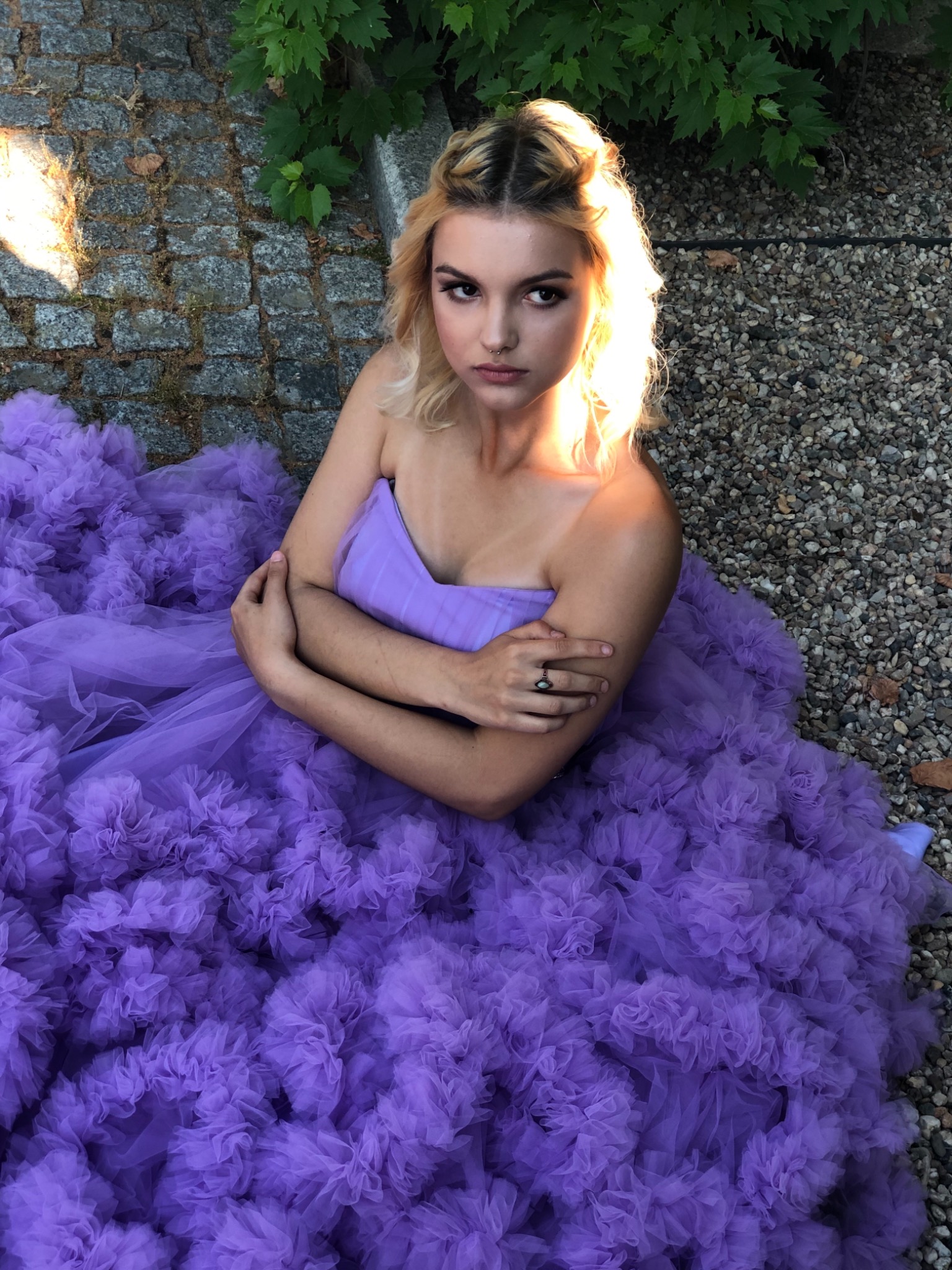 Portret kobiety w fioletowej sukni z falbanami, siedzącej na żwirowym podłożu, oświetlonej słońcem, z delikatnym makijażem i upiętymi włosami.