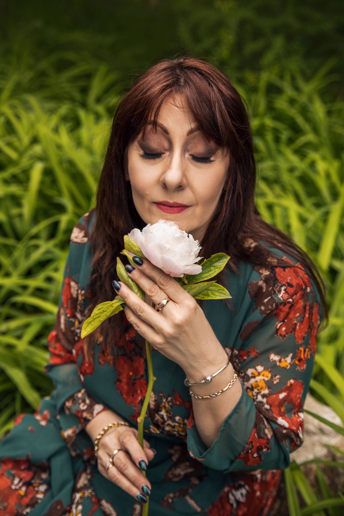 Kobieta z ciemnymi włosami i ciemnymi paznokciami trzyma białą piwonię, zamknięte oczy, zielone tło, złota biżuteria.