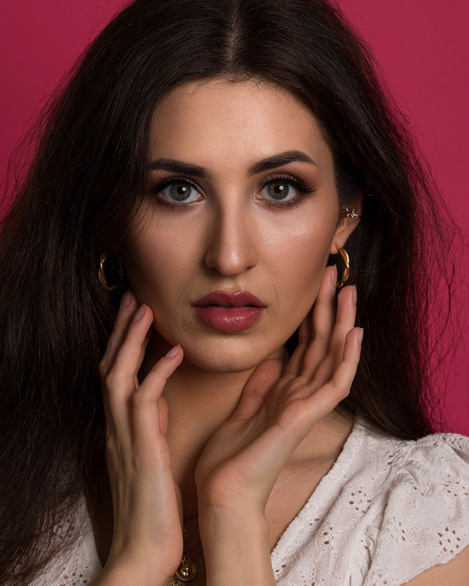 Portret kobiety z długimi, ciemnymi włosami, delikatnym upiększeniem i złotymi kolczykami na tle fuksjowym, dłońmi dotyka twarzy, podkreślając subtelny, dzienny wygląd.