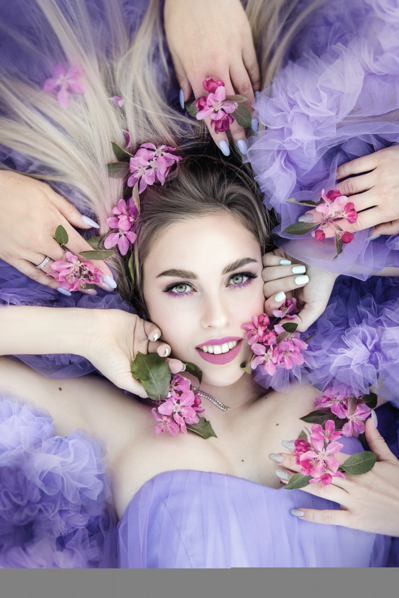 Portret kobiety w fioletowej sukni otoczonej dłońmi z kwiatami jabłoni, makijaż w odcieniach fioletu, platynowe włosy rozłożone wokół głowy.