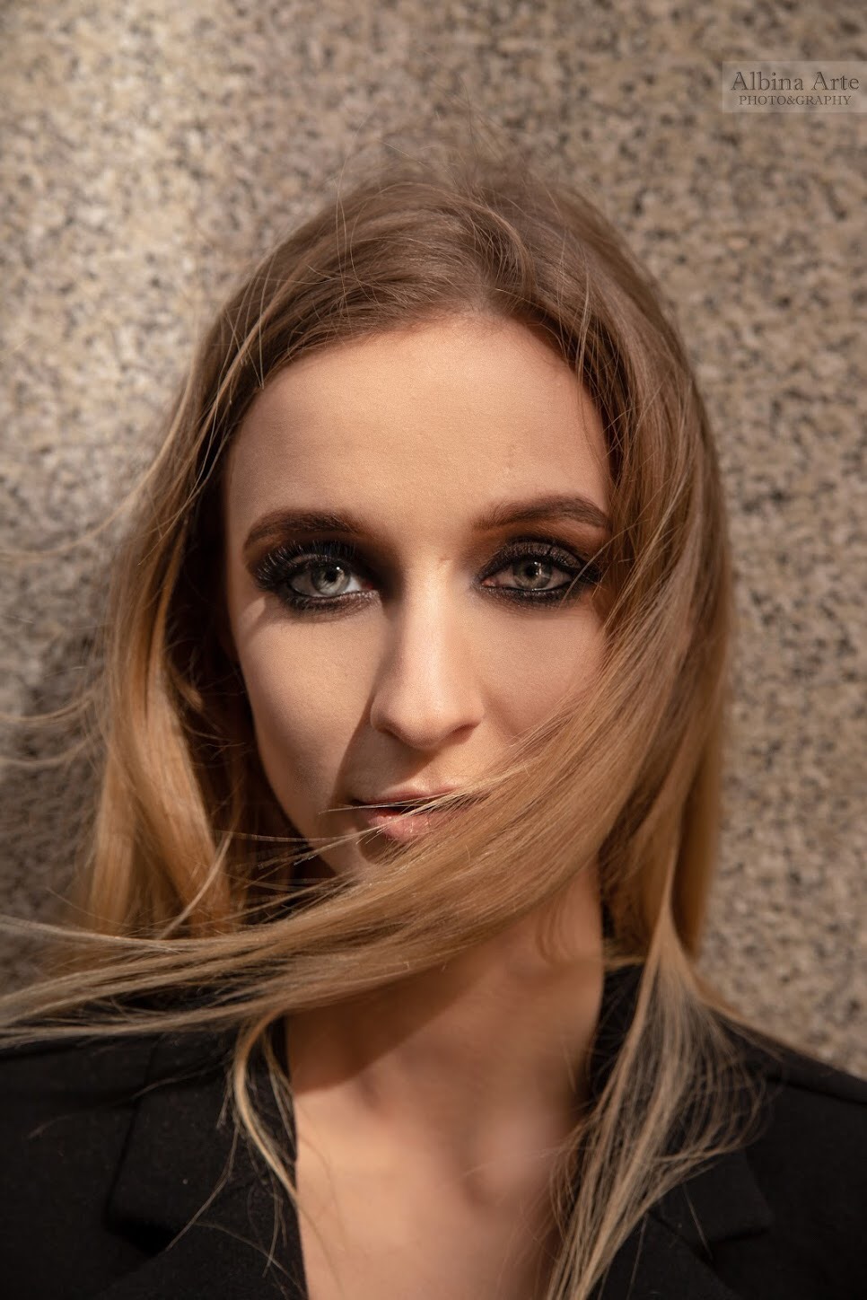 Portret młodej kobiety z intensywnym makijażem oczu w stylu smoky eye, z blond włosami delikatnie rozwianymi na twarzy, na tle szarej, teksturowanej ściany.