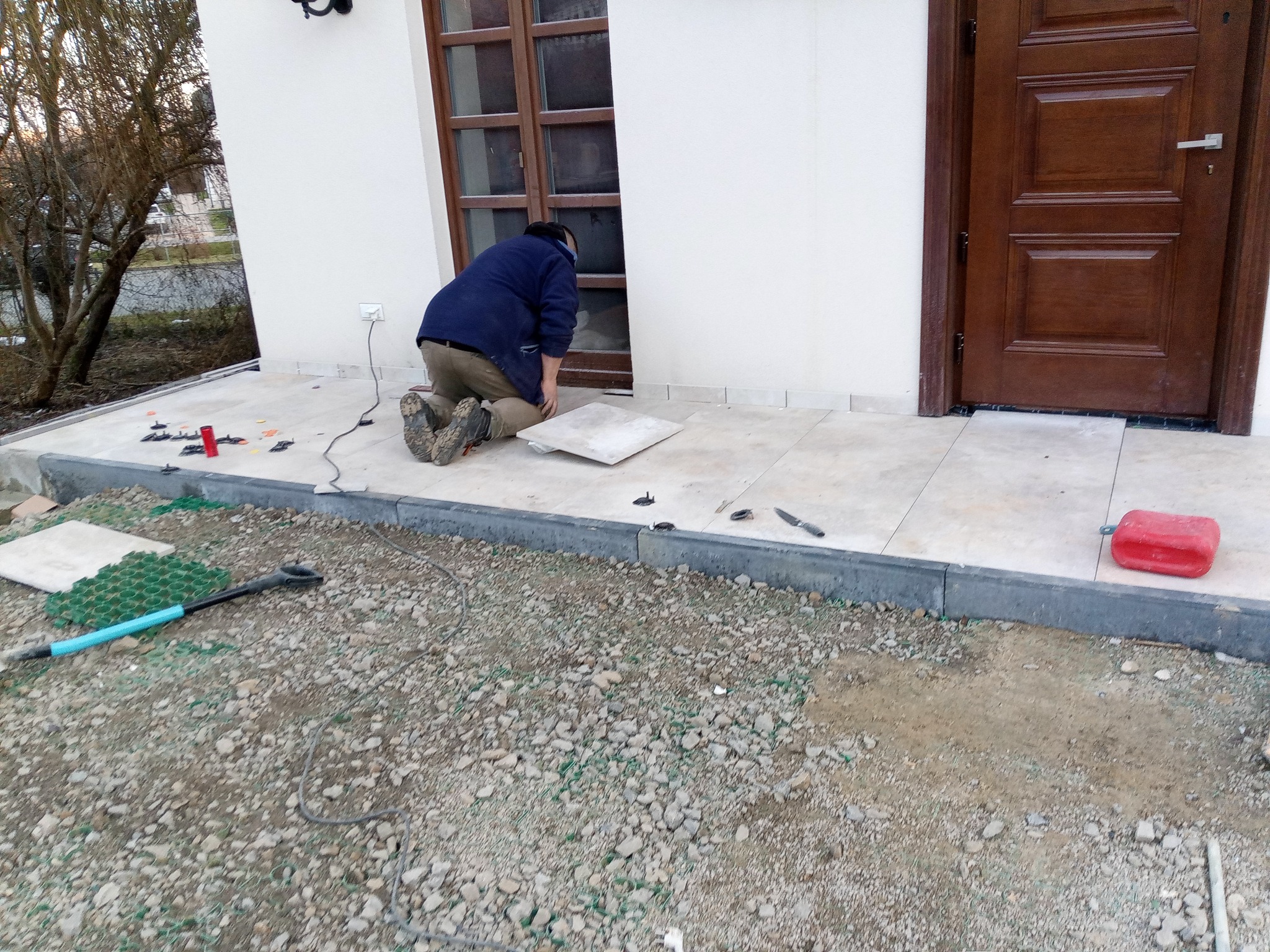 Pracownik na kolanach układa płytki na tarasie przed budynkiem, widoczne narzędzia i materiały budowlane, w tle drzwi wejściowe i okno.