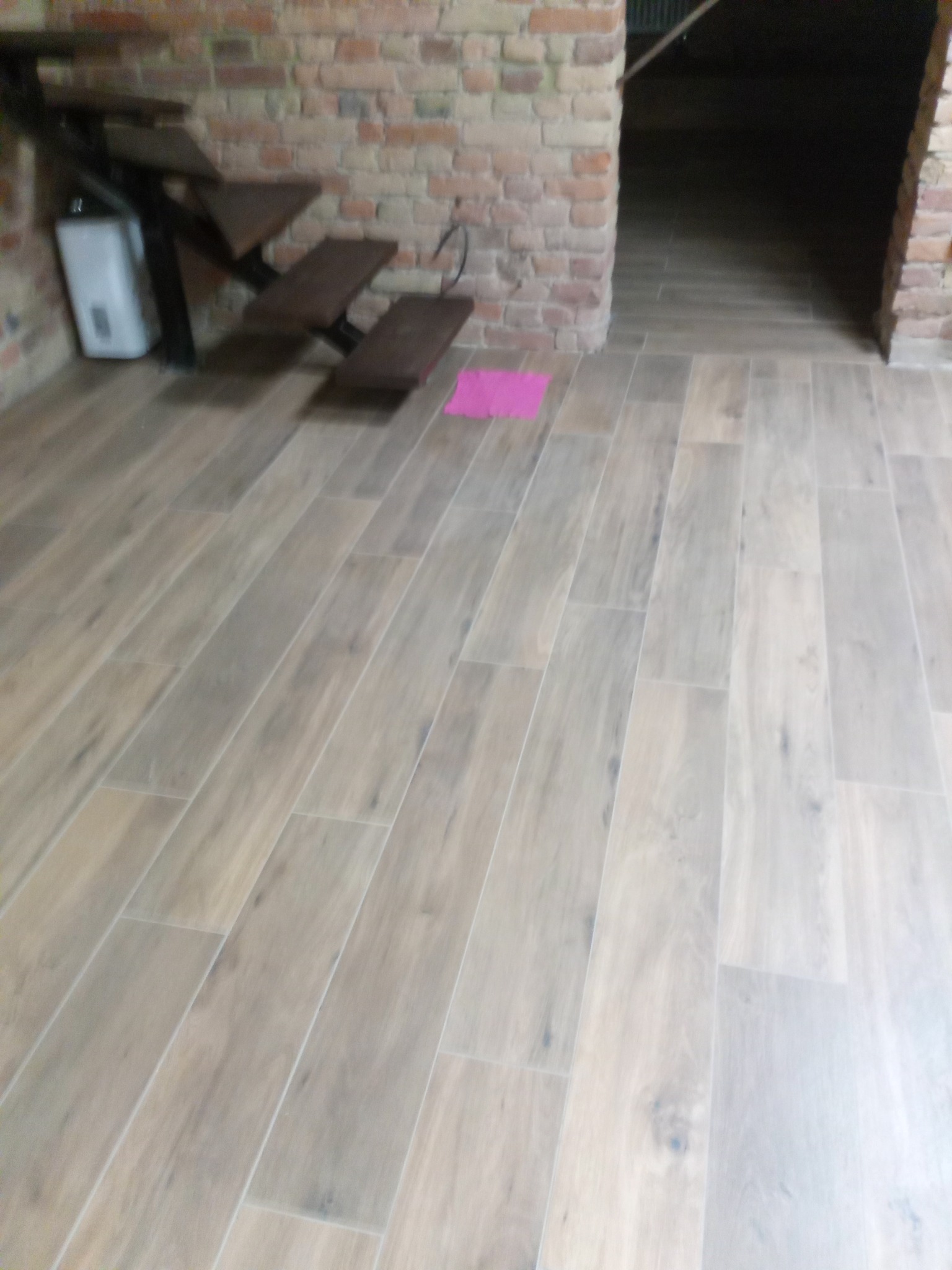 Jasne płytki podłogowe imitujące drewno ułożone wzdłużnie, widoczne schody z ciemnego drewna i fragment ściany z cegły, na podłodze leży różowa ściereczka.