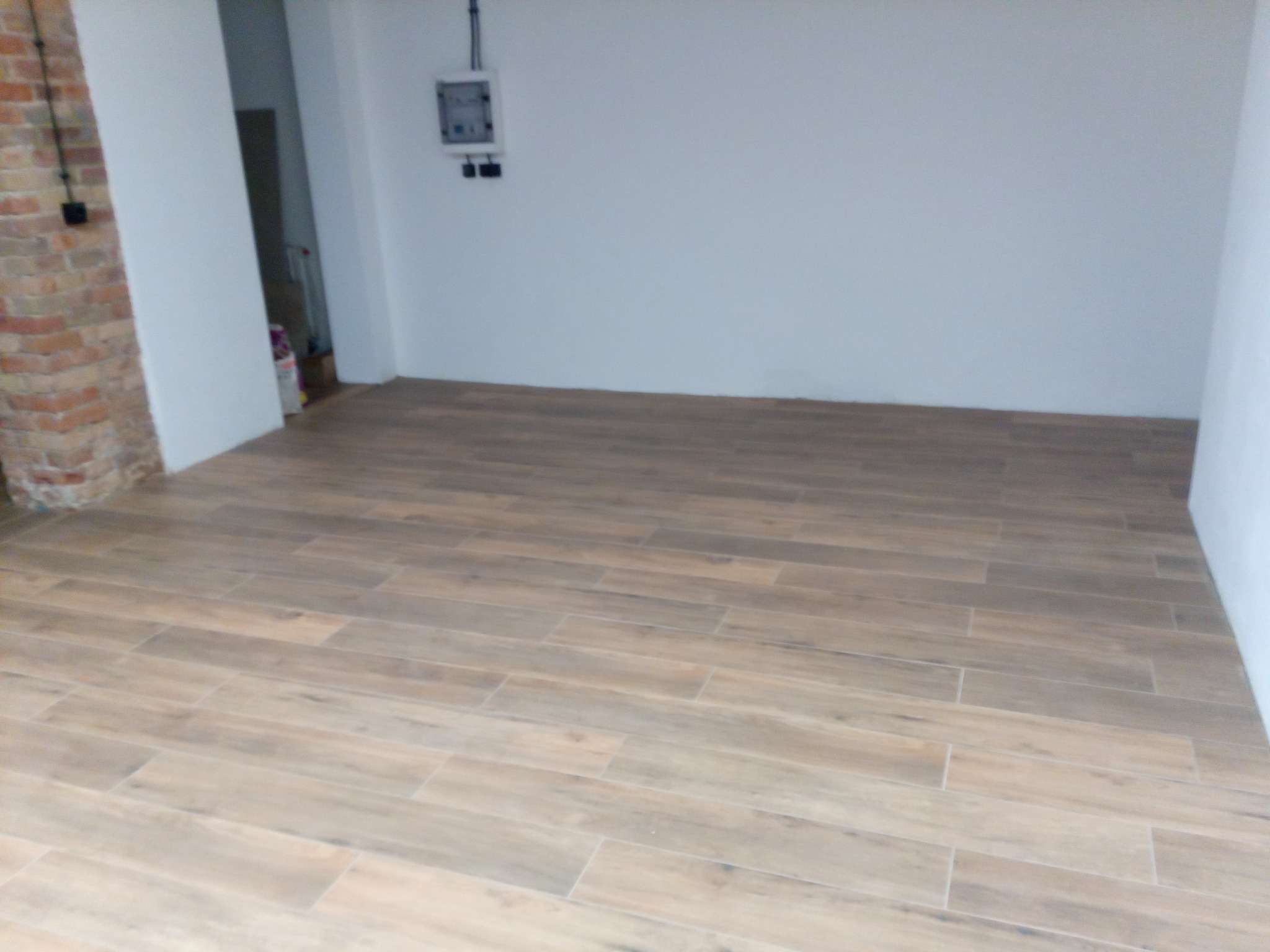 Ułożona podłoga z płytek imitujących drewno w pustym, jasnym pomieszczeniu z fragmentem ściany z cegły i instalacją elektryczną na ścianie.
