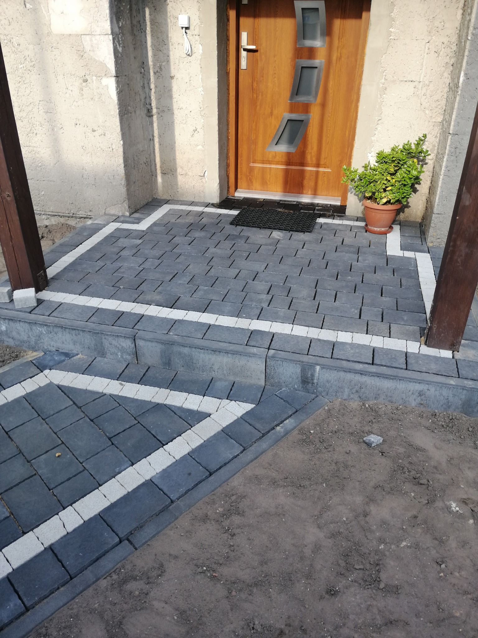 Ułożony chodnik z ciemnoszarej kostki z białymi akcentami przed wejściem do domu, widoczny stopień i donica z rośliną przy drzwiach.