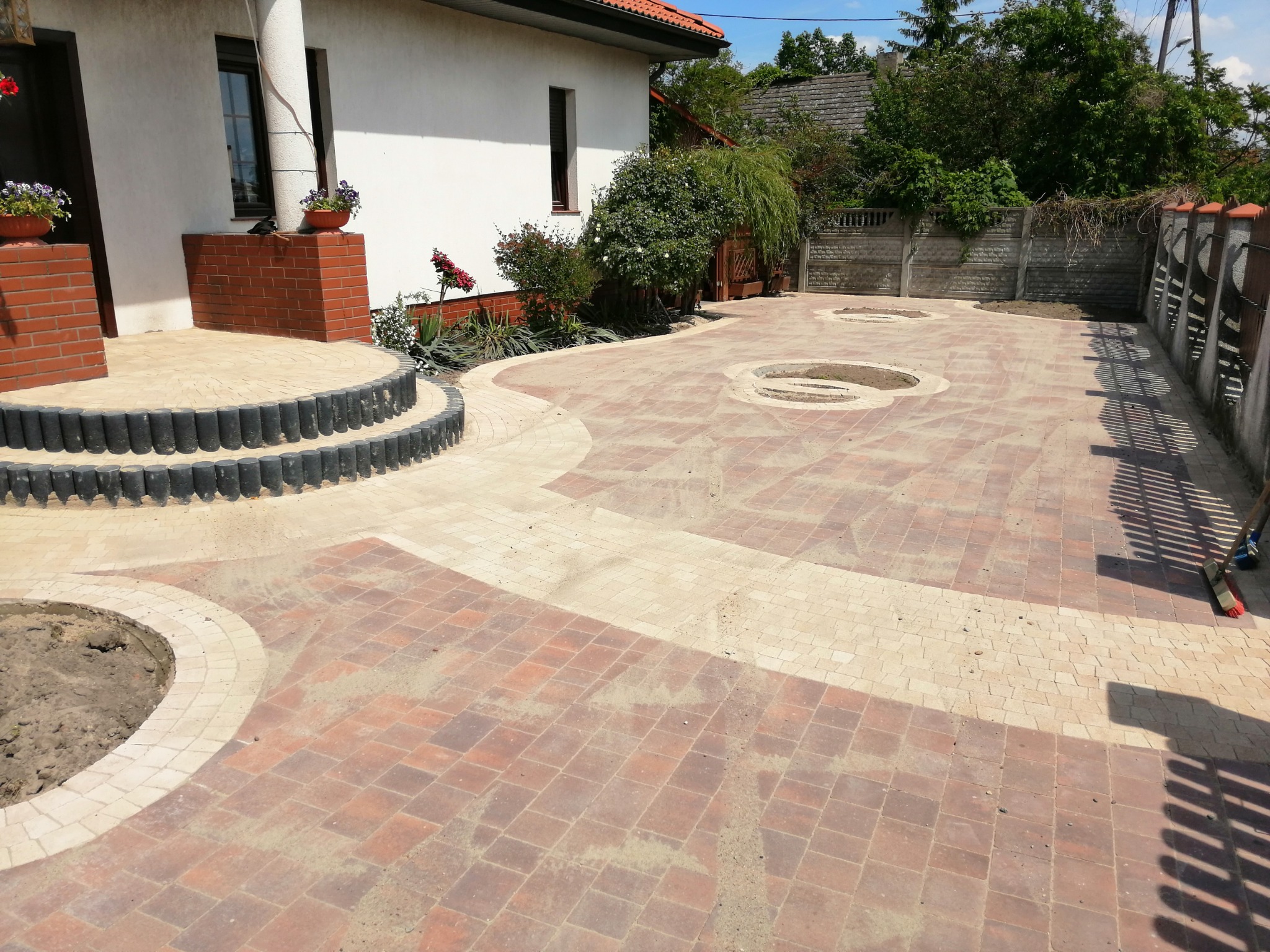 Ułożona kostka brukowa w odcieniach czerwieni i beżu tworząca wzór na podjeździe, z trzema okrągłymi wykopami przygotowanymi pod nasadzenia, widok na dom z elementami ceglanymi i ogród