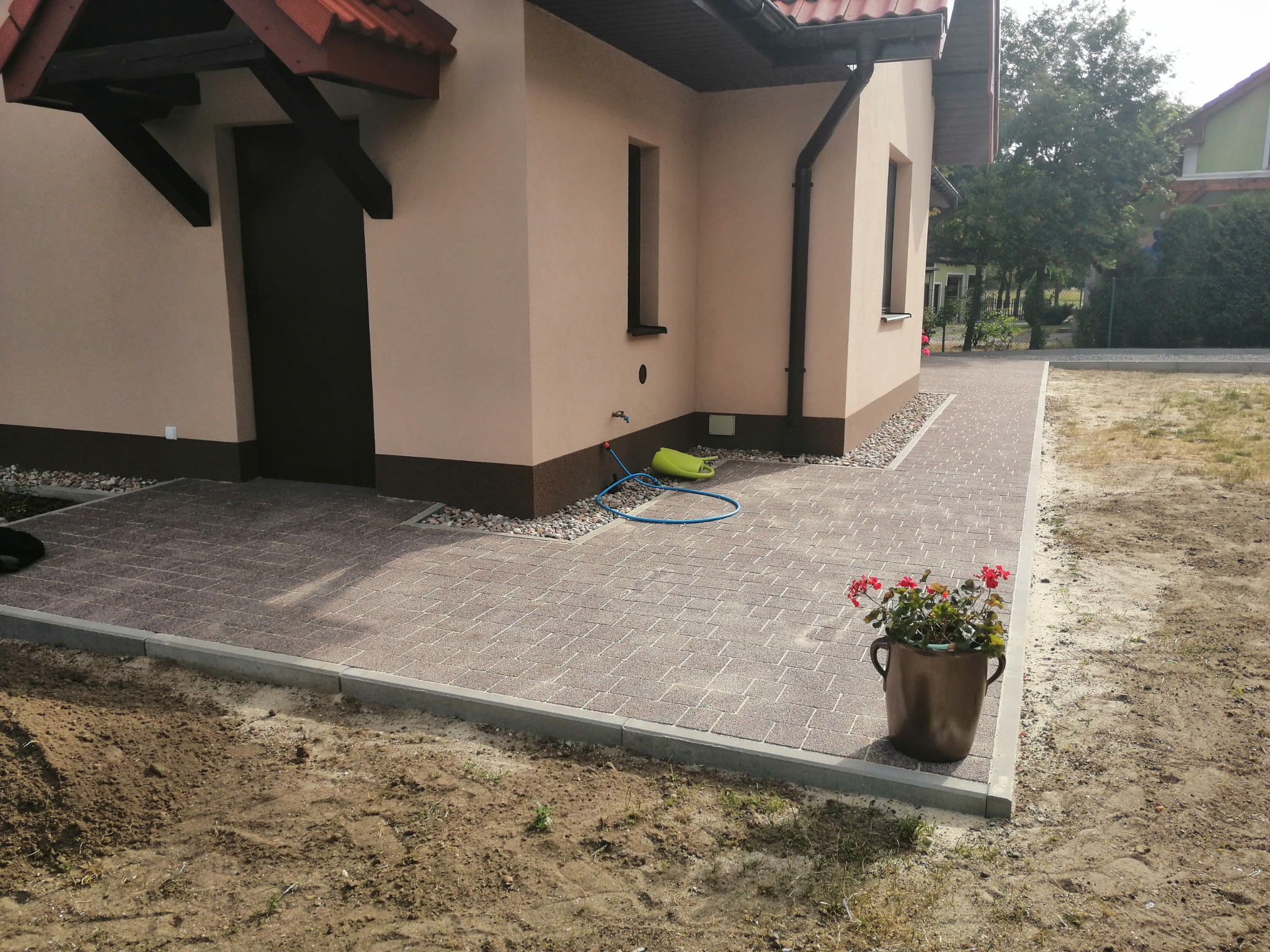 Ułożona kostka brukowa w odcieniach brązu przed domem z beżową elewacją, widoczny wąż ogrodowy i donica z czerwonymi kwiatami.