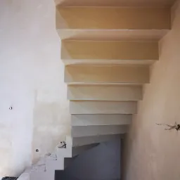 Surowe, otynkowane schody wewnętrzne w trakcie prac wykończeniowych, widoczne niedoskonałości na ścianach i podłodze, z bliska