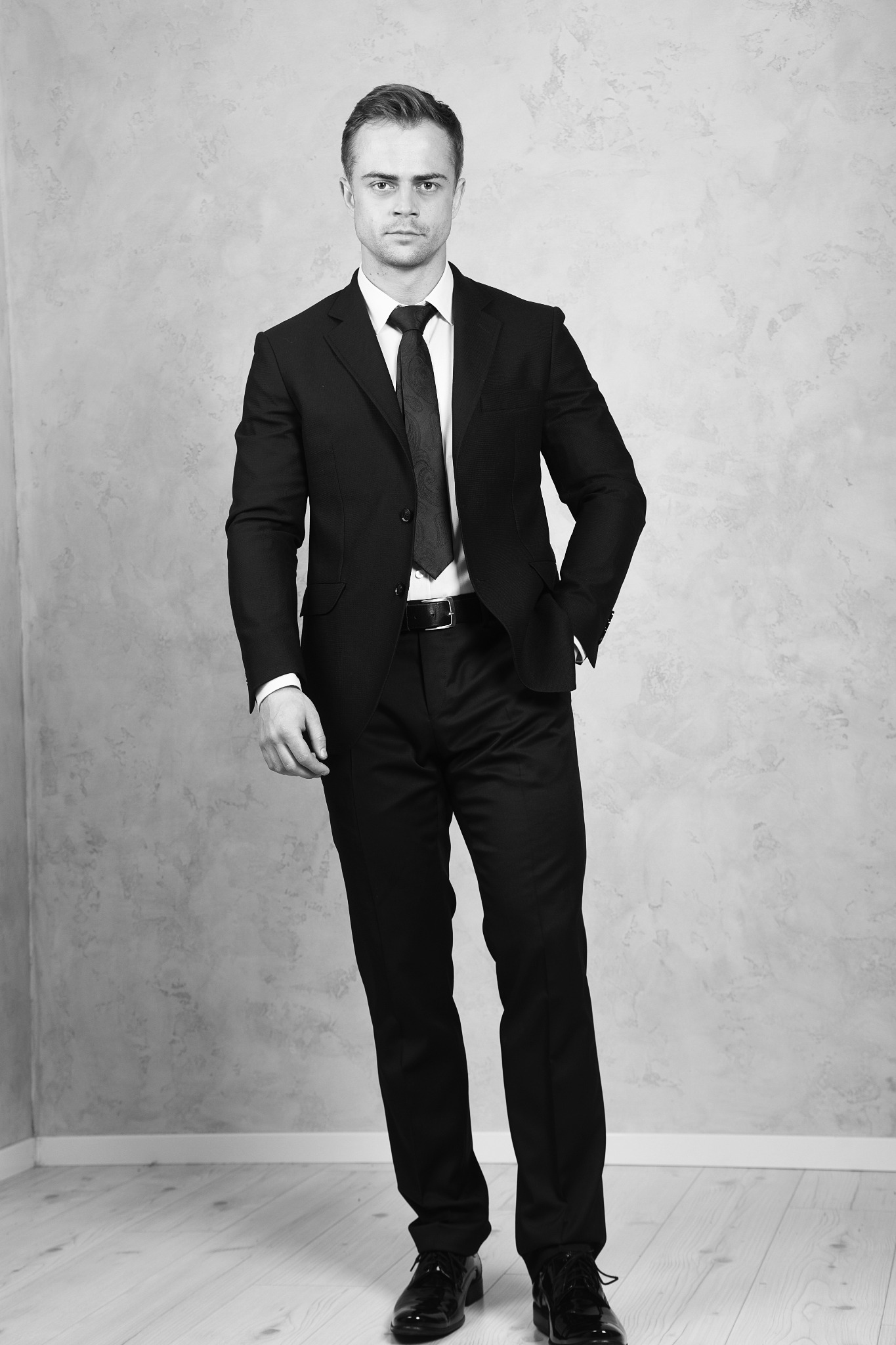 Portret biznesowy mężczyzny w czarnym garniturze i krawacie, stojącego na tle szarej ściany w studio, z jedną ręką w kieszeni.