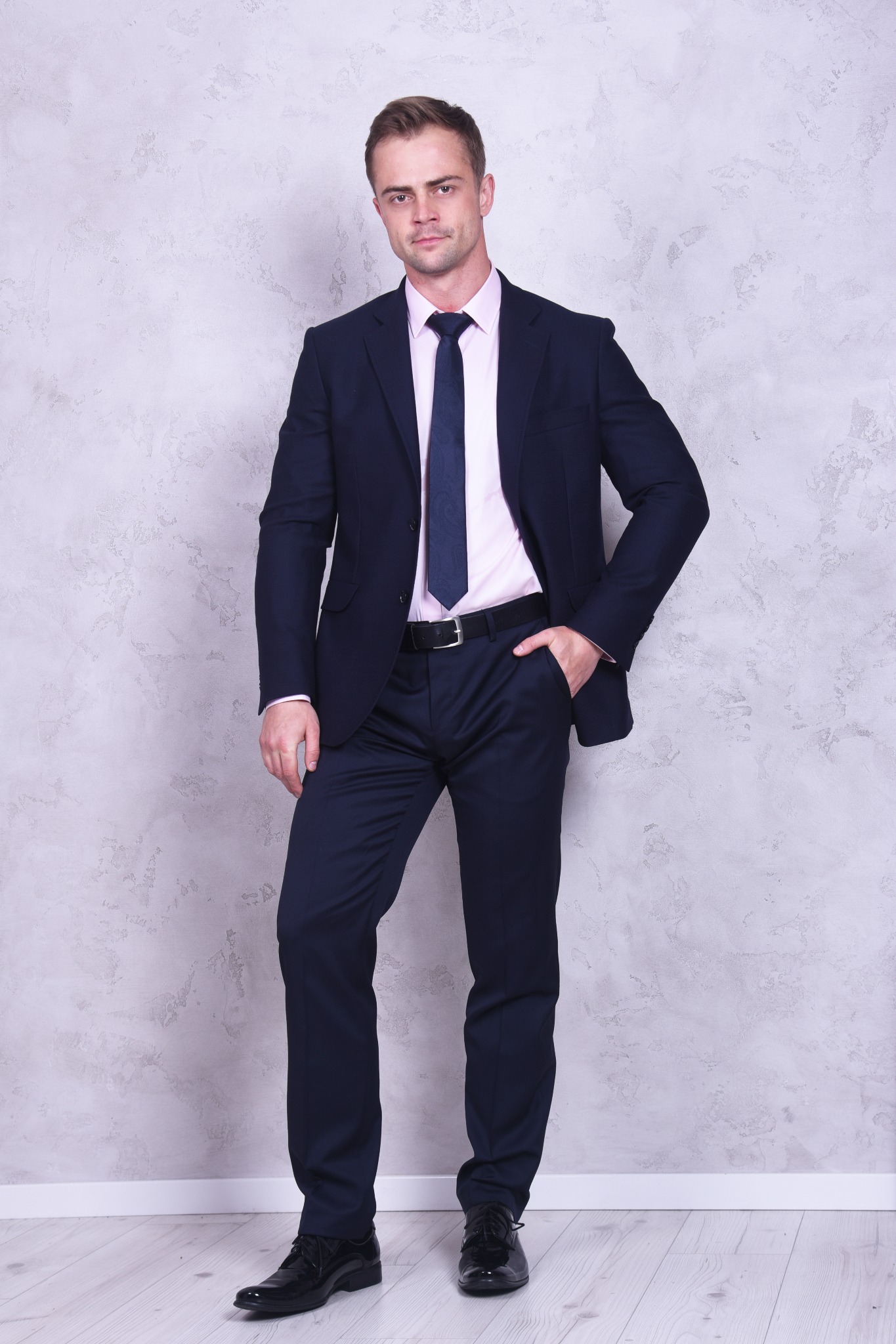 Mężczyzna w granatowym garniturze, różowej koszuli i ciemnym krawacie pozuje na tle szarej, teksturowanej ściany. Ręka w kieszeni spodni.