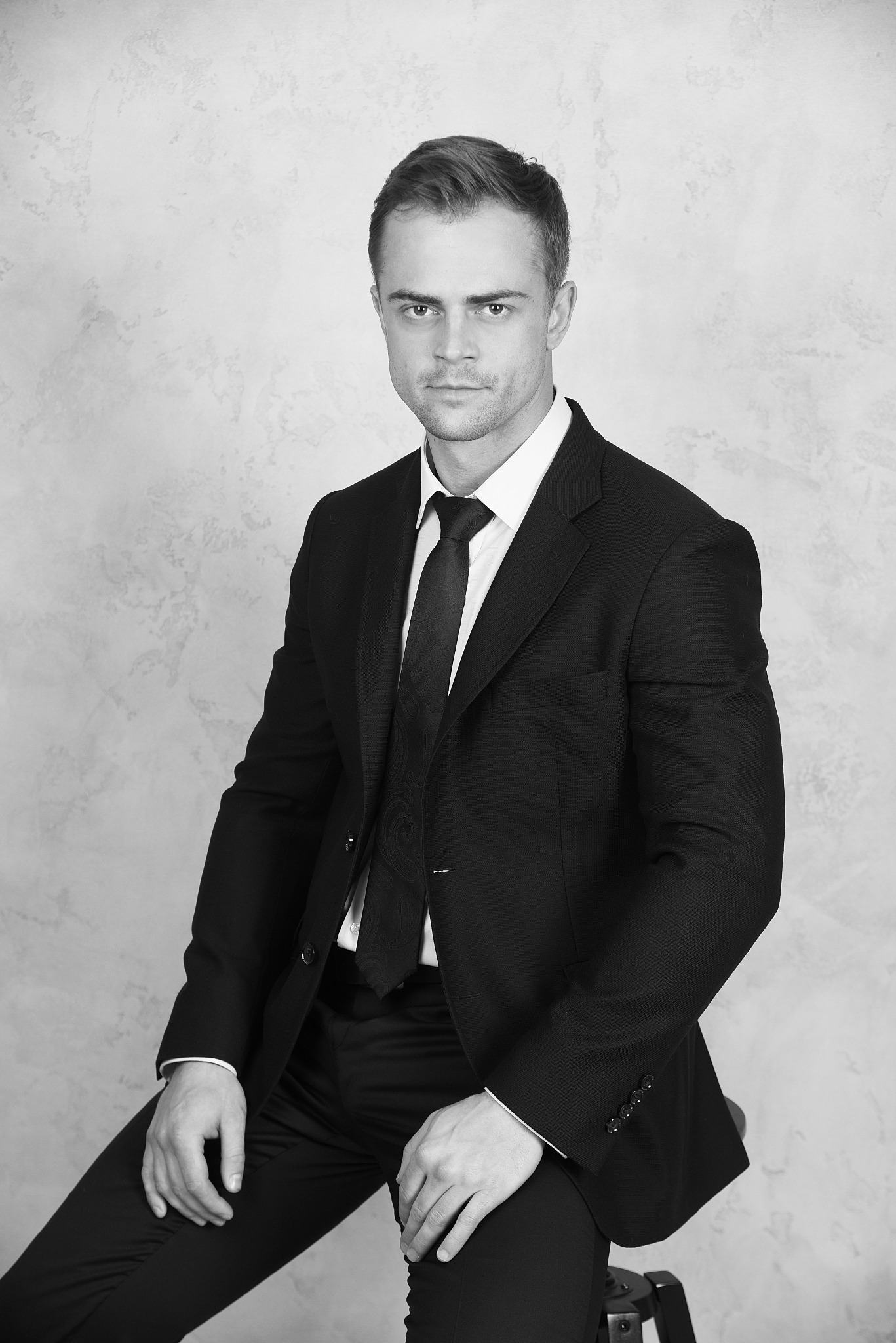 Portret mężczyzny w czarnym garniturze, siedzącego na stołku na tle szarej ściany o teksturze betonu, z subtelnym wzorem na krawacie, w czerni i bieli.