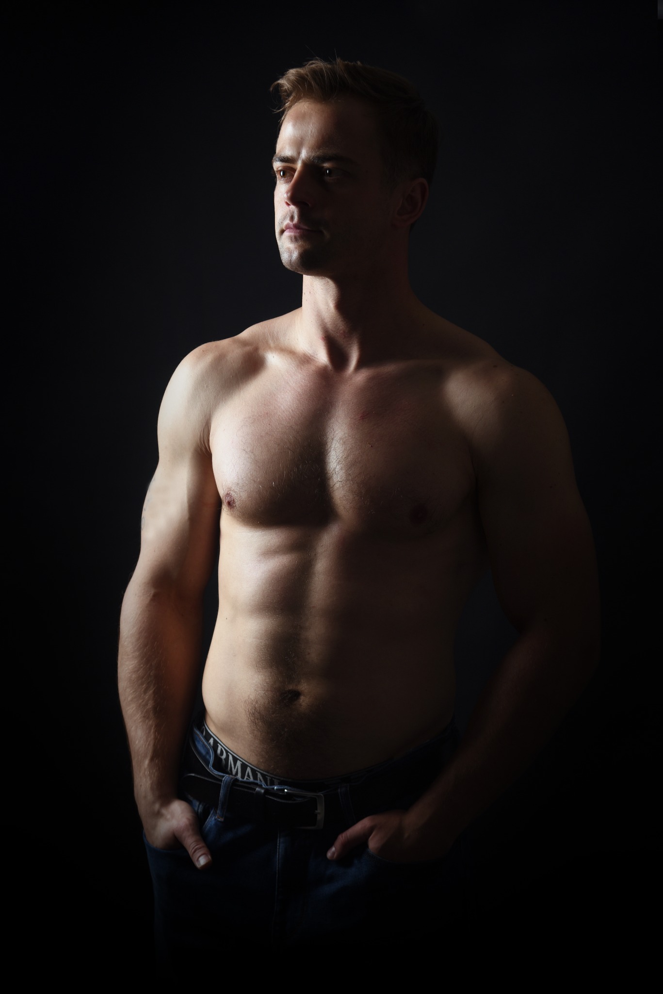 Mężczyzna o atletycznej budowie ciała, częściowo obnażony, pozuje w niebieskich jeansach na ciemnym tle, oświetlony z boku, z rękami w kieszeniach.