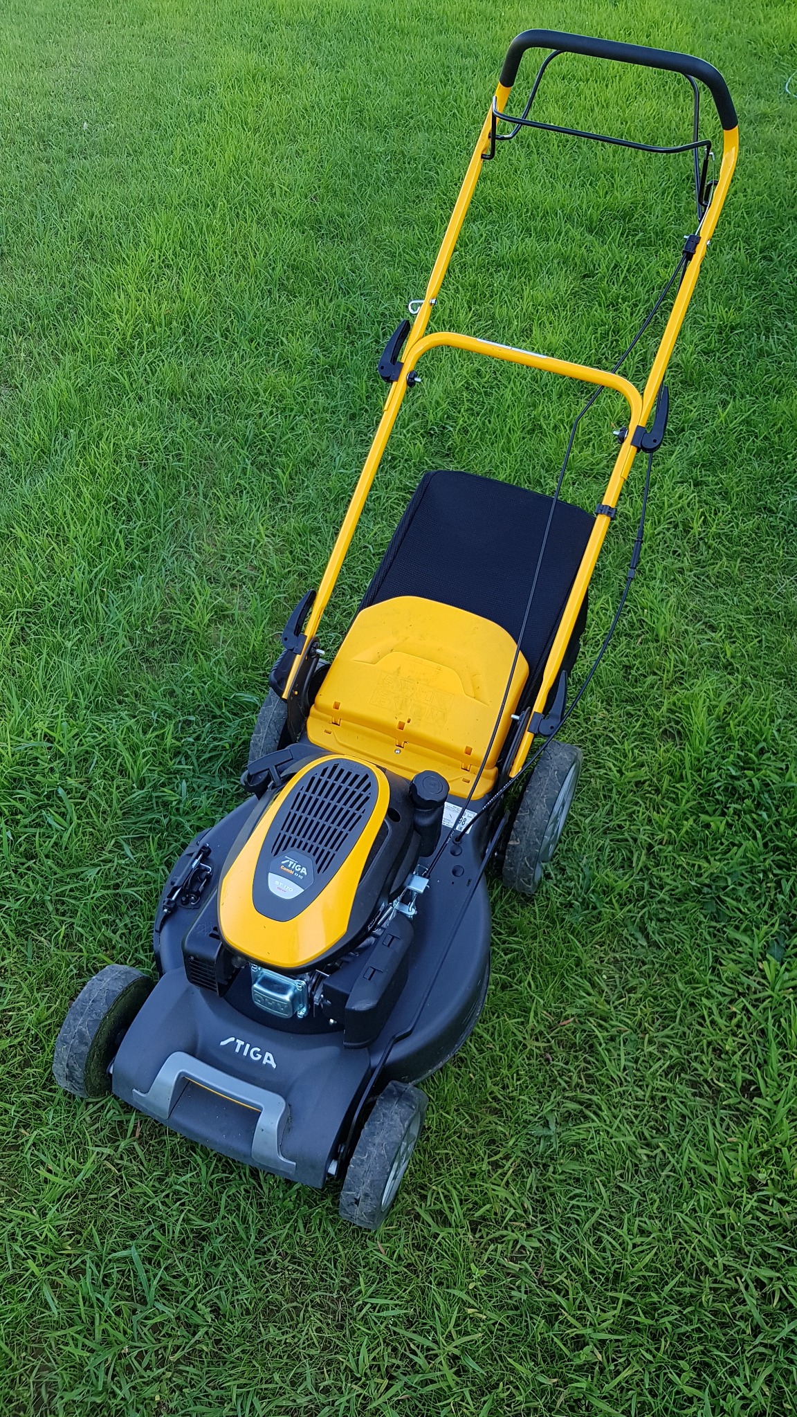 Żółto-czarna kosiarka spalinowa marki Stiga stoi na trawniku z widocznymi śladami użytkowania na kołach.