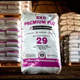 Worek ekogroszku EKO PREMIUM PLUS o wadze 25 kg, z widocznymi informacjami o granulacji, wartości opałowej i zawartości popiołu, ustawiony na tle palet z zapasem worków w magazynie.