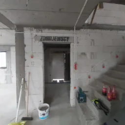 Wnętrze w stanie surowym z widoczną instalacją elektryczną poprowadzoną po ścianach, kable w białych listwach z czerwonymi puszkami, schody betonowe, wiadro po farbie i narzędzia na podłodze.