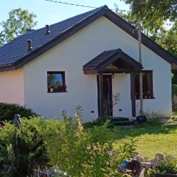 Gotowy, parterowy dom z białymi ścianami, ciemną dachówką i drewnianym zadaszeniem nad wejściem, otoczony zielonym ogrodem z kwiatami w doniczkach na parapetach.