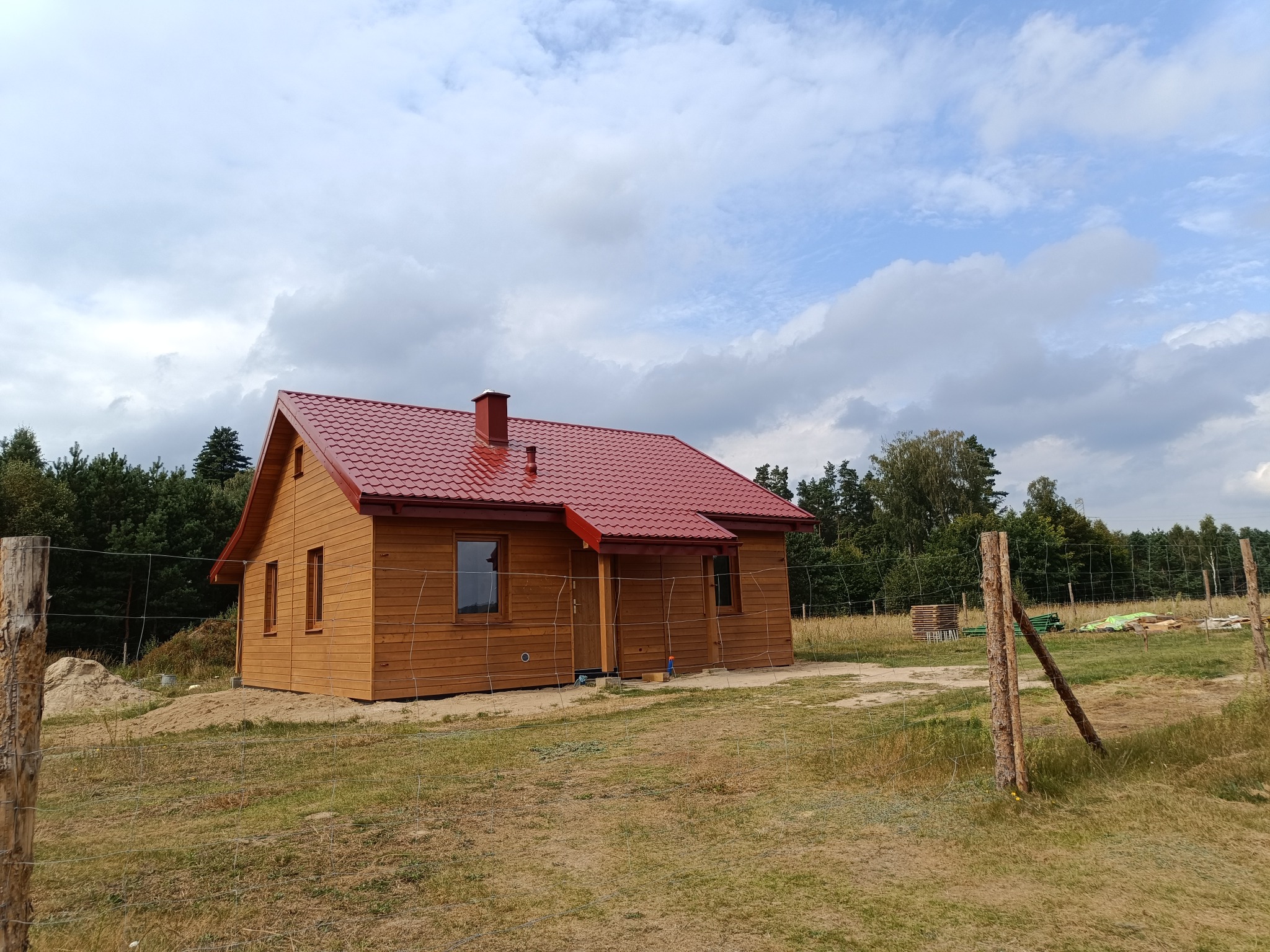 Gotowy, parterowy dom z drewnianych bali z czerwoną dachówką, widoczny przez ogrodzenie z drewnianych słupków i siatki.
