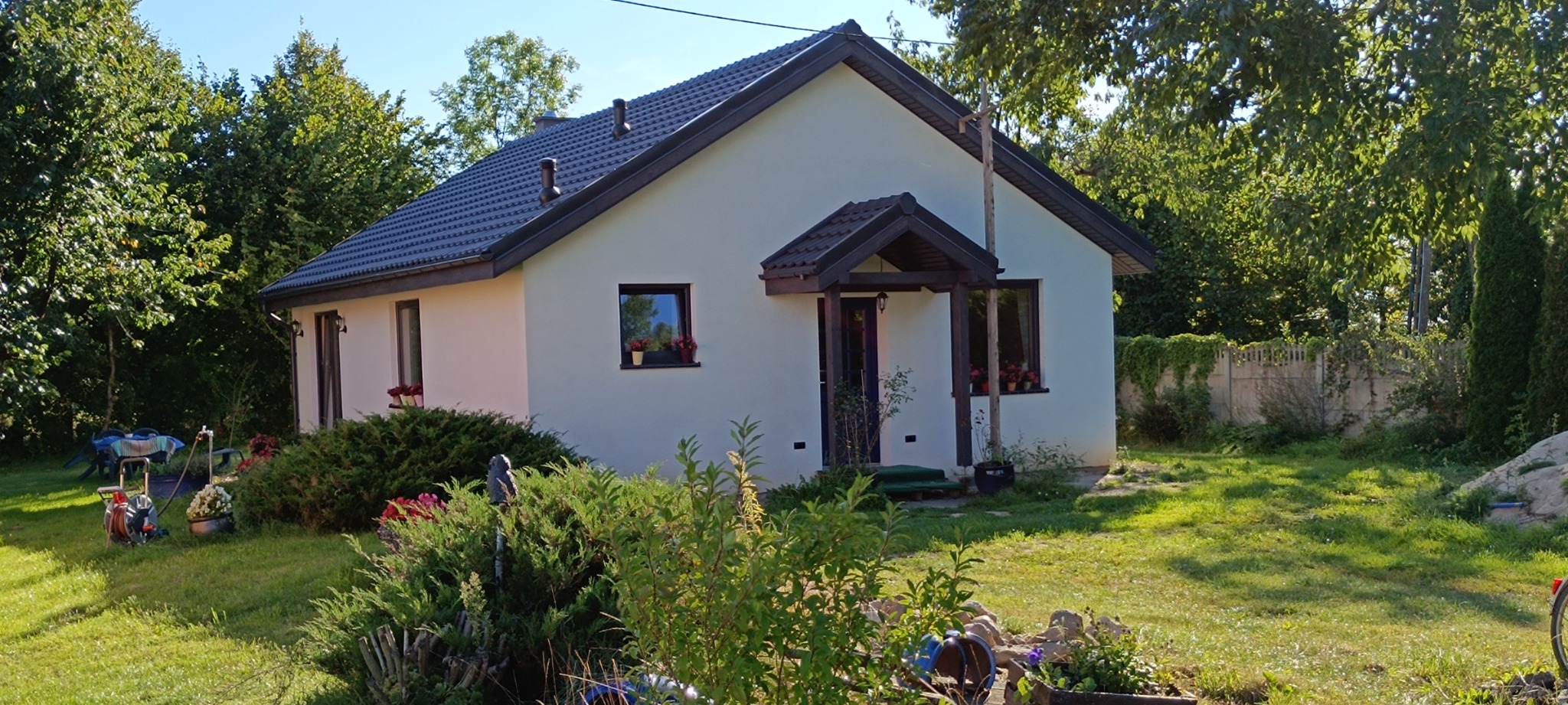 Gotowy, parterowy dom z białymi ścianami, ciemną dachówką i drewnianym zadaszeniem nad wejściem, otoczony zielonym ogrodem z kwiatami w doniczkach na parapetach.