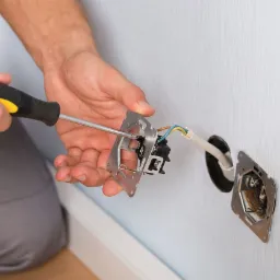 Elektryk przykręca metalową obudowę gniazdka elektrycznego do ściany za pomocą śrubokręta z czarno-żółtą rączką, widoczne przewody w ścianie.