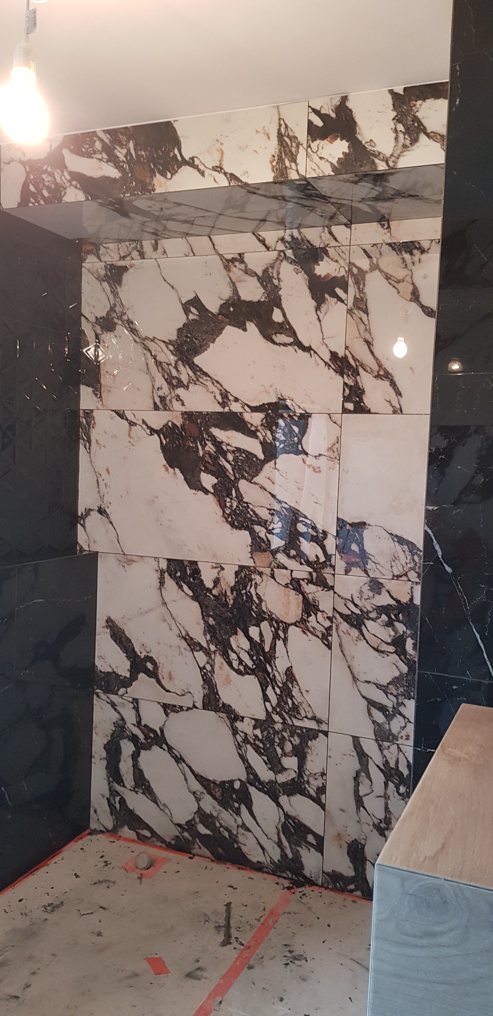 Ściana łazienki wyłożona płytkami imitującymi marmur w odcieniach bieli i czerni, kontrastująca z ciemnymi, fakturowanymi płytkami po bokach; widoczna surowa podłoga z zaznaczonymi taśmą granicami,...